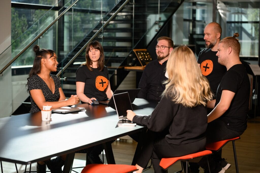 Six people are seated around a rectangular table in an office setting. Five are facing a woman on the left who is speaking. Laptops and a coffee cup are on the table. Some people wear matching black shirts with an orange circle and a black cross on them. A staircase is in the background.