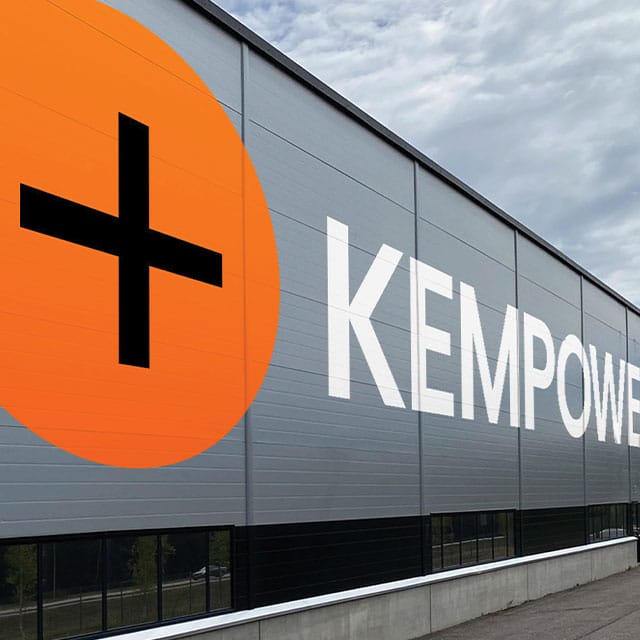 A large grey industrial building with the word "KEMPOWER" prominently displayed on the side in white capital letters. To the left of the text, there is an orange circle with a black plus sign inside it. The sky above is partly cloudy.