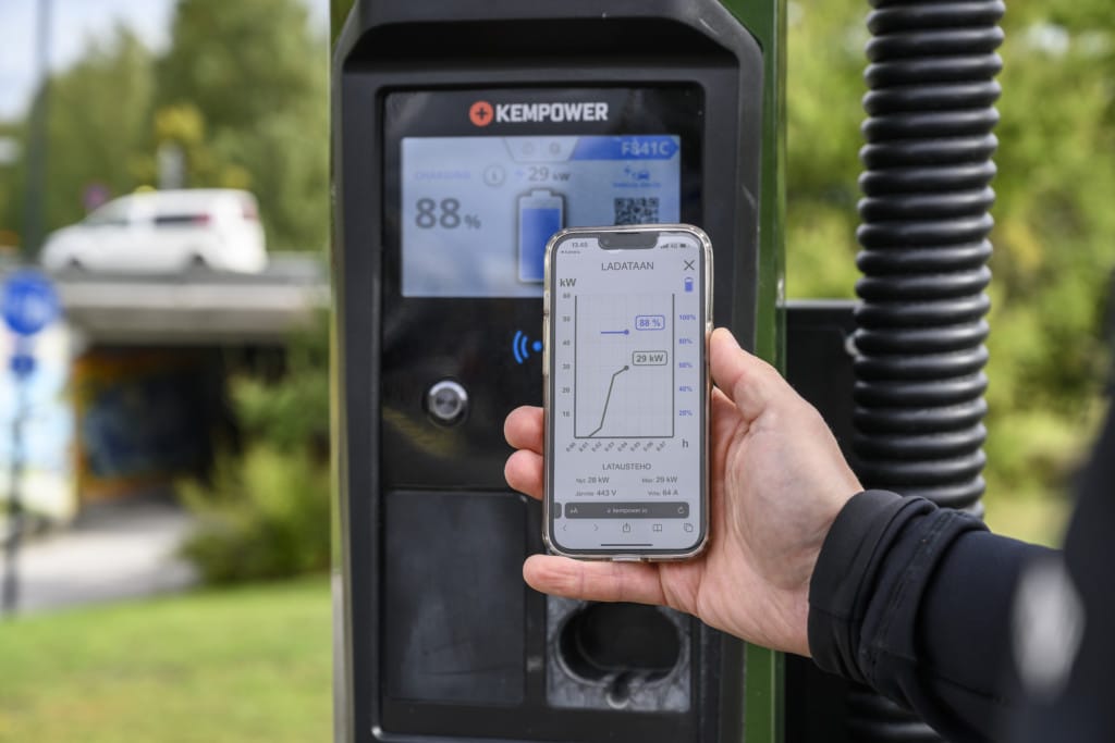 A person holds a smartphone displaying a charging graph in front of a Kempower electric vehicle charging station.
