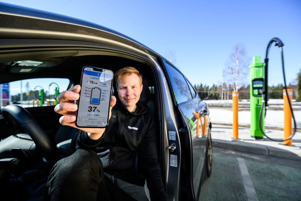 A person sitting in a car holds up a smartphone displaying a charging app, with electric vehicle charging stations visible outside.