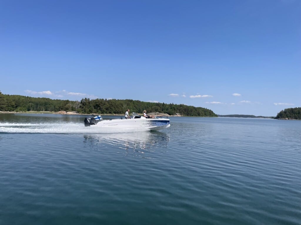 A speedboat glides across a calm, blue lake under a clear sky with distant trees lining the shore.