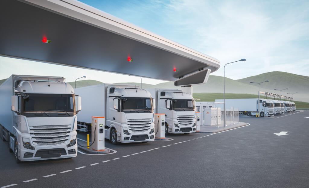 Three white trucks are lined up at an outdoor charging station under a modern, wide canopy against a backdrop of green hills and a clear blue sky.