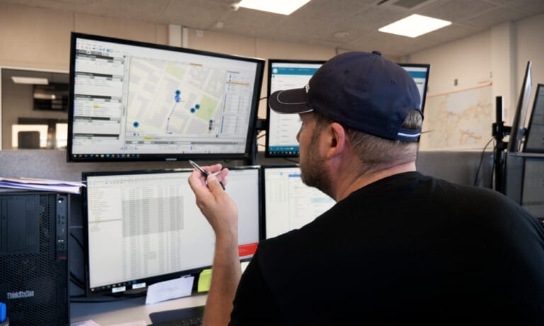 A man wearing a cap sits at a desk with multiple monitors displaying maps and data, holding a stylus pen, possibly analyzing information or managing a control system.