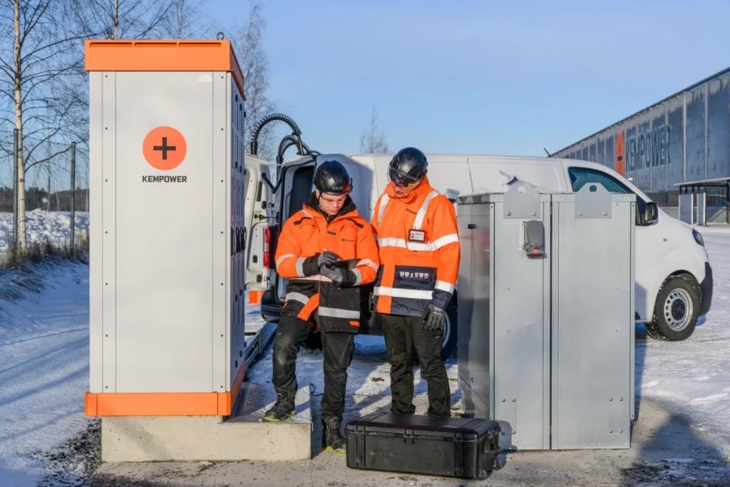 Two technicians in high-visibility jackets work on an electrical charging station outdoors in a snowy environment, using a digital device. an electric van is parked in the background.