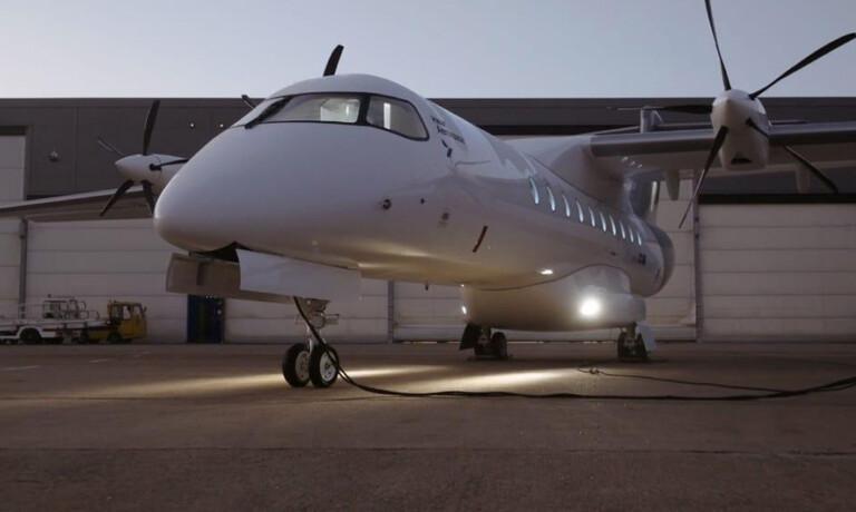 A white twin-propeller airplane is parked on the tarmac in front of a hangar at dusk, with lights illuminating the ground beneath it.